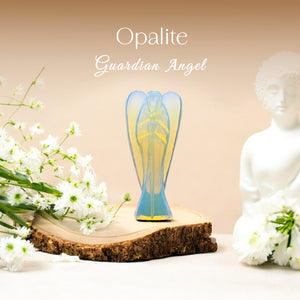 An Opalite Guardian Crystal Angel figurine is displayed on a wooden surface with white flowers around it. The angel figurine has a soft, translucent blue color with patterns that resemble a guardian angel.